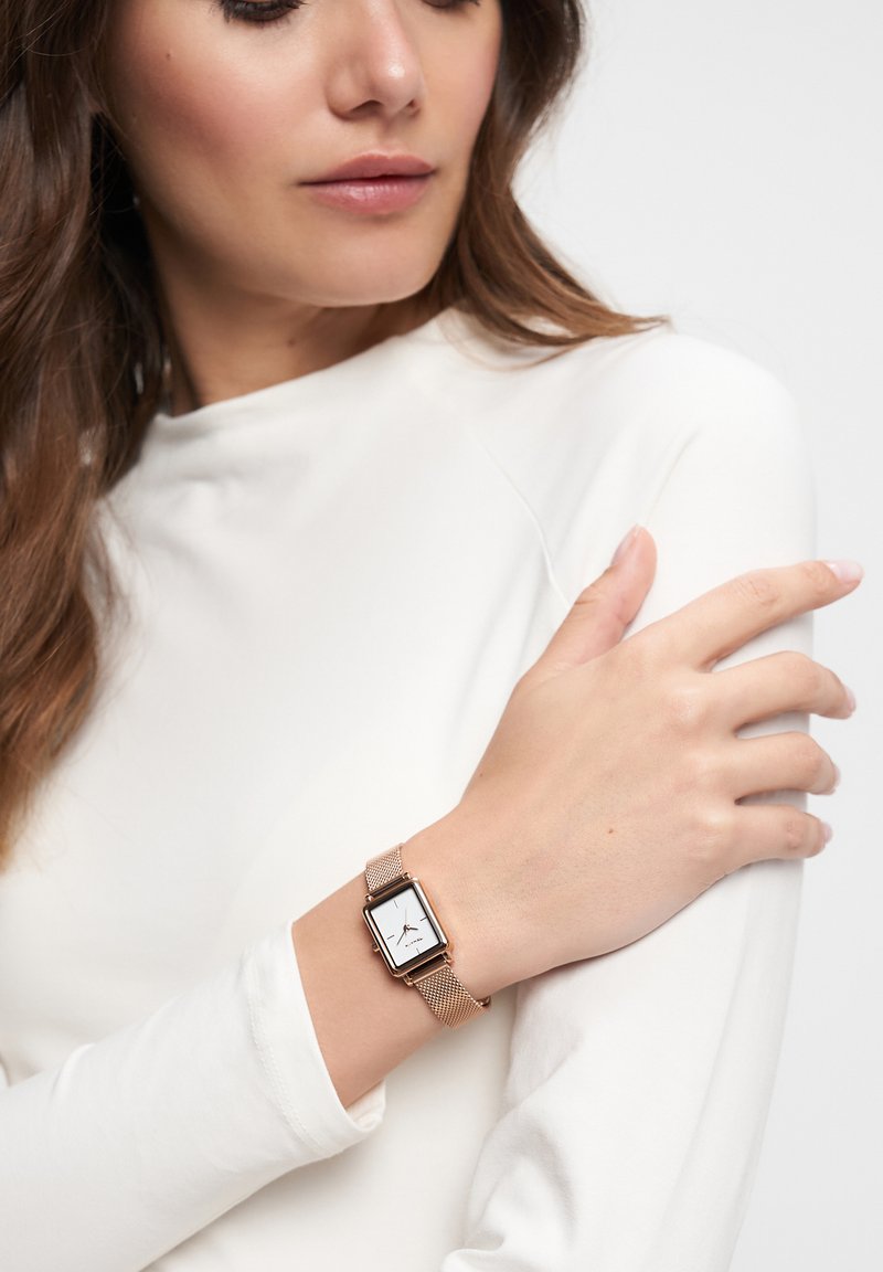 Roségoldenes Mesh-Armbanduhr mit rechteckigem Ziffernblatt, weißem Zifferblatt und schwarzen Stundenmarkierungen, getragen an einem Handgelenk mit einem glatten, figurbetonten weißen Oberteil.