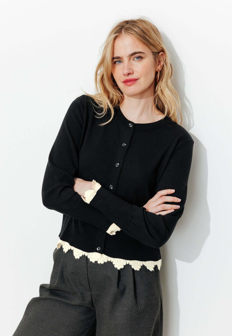 Femme blonde portant un cardigan noir boutonné avec des bordures en dentelle blanche et un pantalon gris, se tenant les bras croisés devant un fond blanc.