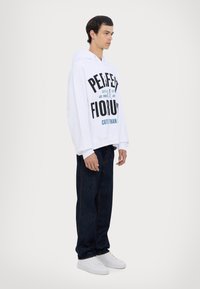 Hvid hættetrøje med fed sort og teal tekst, lagt sammen med mørk denim jeans og hvide sneakers, har en afslappet pasform og lange ærmer.