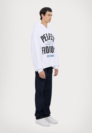 Weißer Hoodie mit auffälligem schwarzen und türkisfarbenem Text, kombiniert mit dunklen Denim-Jeans und weißen Sneakers, im lässigen Schnitt und mit langen Ärmeln.