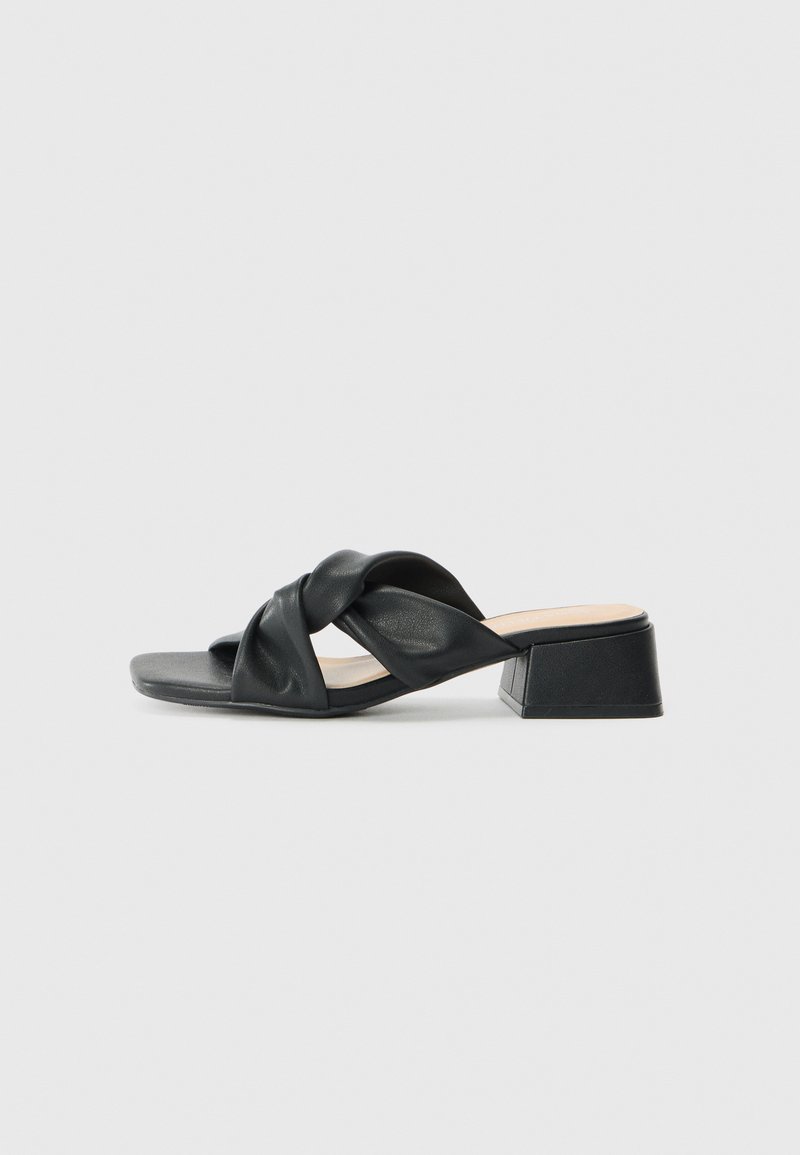 Sandalia de cuero negro con un diseño superior retorcido, punta cuadrada y un bajo tacón bloque, con una textura suave y un estilo minimalista.