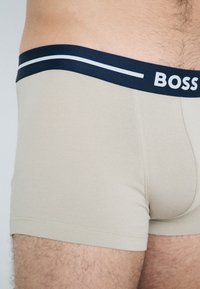 Beige boxernadrág sötétkék derékrésszel, amelyen fehér csíkok és a "BOSS" logó található. Sima textúrájú, testhezálló fazon, közép-comb hosszúságú.