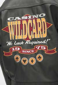 Melna ādas jaka ar krāsainu izšuvumu: "CASINO WILDCARD" sauklis, ar spēļu kāršu mastu dizainiem un gadu "1975".