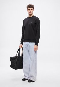 Čierna mikina s okrúhlym výstrihom a malým logom, svetlomodré široké rifle, čierna taška na nosenie a čierne topánky. Hladké textúry a nenápadný dizajn.