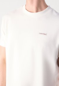 Osoba oblečená v jednoduchém bílém tričku Calvin Klein s malým logem značky na levé hrudi, zobrazená od krku po střed trupu.
