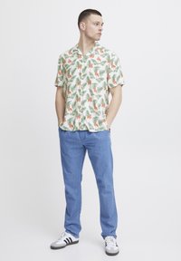 Kurzarmhemd mit weißem Hintergrund, das ein florales Muster in Grün und Orange zeigt. Kombiniert mit hellblauen Jeans und schwarz-weißen Sneakers.