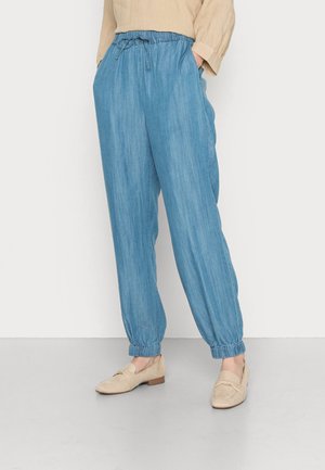 Personne portant un pantalon en denim bleu clair à taille élastique et coupe fuselée, ainsi que des mocassins beiges sans lacets, debout les mains dans les poches devant un fond uni.