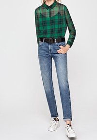 Camisa de manga larga de cuadros verdes y transparente con cuello, cinturón negro, jeans de color azul claro y zapatillas blancas con acentos negros.