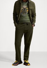 Completo verde oliva con un maglione lavorato a maglia con design di un orso, pantaloni jogger coordinati e sneakers nere con accenti gialli. Tessuto strutturato, vestibilità rilassata.