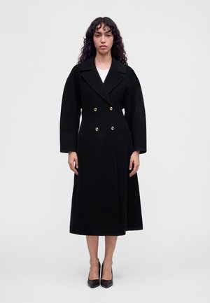 Femme aux longs cheveux bouclés se tenant face à l’avant, portant un long manteau noir croisé avec des boutons dorés et des talons hauts noirs.
