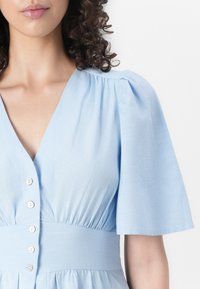 Rochie bleu deschis dintr-un material texturat, cu decolteu în V, mâneci scurte tip puff și nasturi decorativi pe față.
