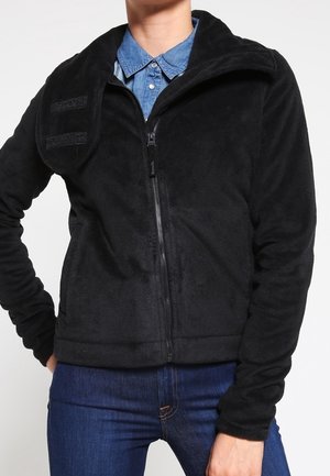 Person iført en sort fleecejakke med lynlås over en blå skjorte med knapper og blå jeans, stående med den ene hånd på hoften.