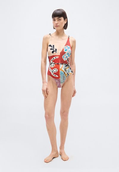 Γυναίκα που στέκεται ξυπόλυτη φορώντας ένα πολύχρωμο, μοτίβο ολόσωμο μαγιό με λεπτές τιράντες και ζώνη στη μέση, μπροστά σε απλό φόντο.