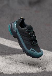 Adidas Terrex fekete és kék túracipő, fűzős és robusztus talppal, aszfalt felett felfüggesztve, fehér útjelzéssel.
