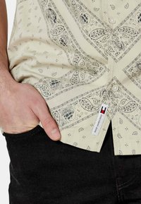 Camicia beige con bottoni, decorata con stampe paisley nere e motivi nautici, con orlo curvo e etichetta del marchio Tommy Hilfiger vicino alla cucitura.
