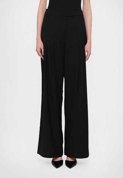 Pantalones de pierna ancha negros, confeccionados en una tela suave, con una cintura alta entallada y sutiles pliegues, combinados con tacones negros de punta.