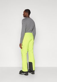 Άνδρας που κοιτάει sideways, φορώντας γκρι μακρυμάνικη μπλούζα, φωτεινά κίτρινα παντελόνια σκι και μαύρες μπότες σκι με την επωνυμία "HEAD".