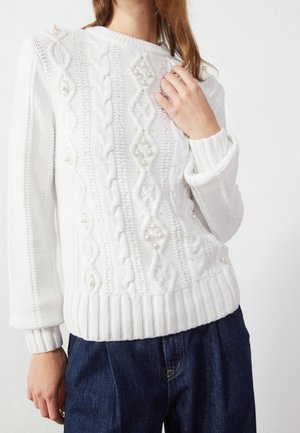 Femme portant un pull en maille torsadée blanc avec des accents en perles et un jean bleu foncé, main levée vers la poitrine.