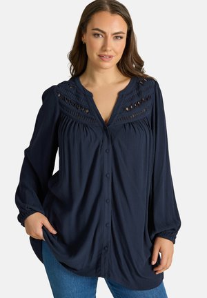 Femme aux longs cheveux bruns portant un chemisier bleu marine à boutons avec des détails en dentelle et des manches longues, associé à un jean bleu.