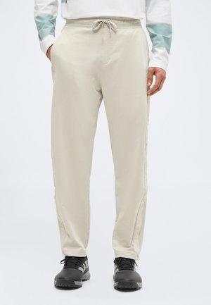 Személy bézs színű, oldalt patentokkal ellátott húzózsinóros nadrágban és fekete sportcipőben, zsebeibe dugott kézzel áll.