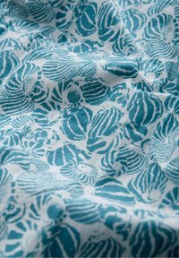 Biała tkanina z niebiesko-zielonymi wzorami muszelek, ukazana z naturalnymi fałdami i miękką teksturą.