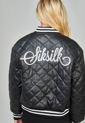 Personne blonde portant une veste matelassée noire avec des poignets et un col à rayures blanches, avec "Siksilk" brodé en blanc dans le dos.
