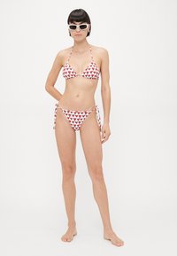 Red heart-patterned bikini made of stretchy fabric; triangle top with halter neck, side-tied bottoms. Model stands barefoot, wearing sunglasses.