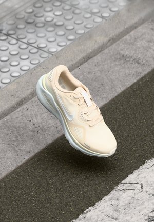 Chaussure de sport beige clair avec tige en mesh, semelle en caoutchouc et accents blancs. Présente un bout arrondi, des lacets et un logo sur le côté.
