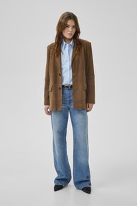 Sieviete, kas valkā br ūnu žaketi, gaiši zilu pogājamās krekla kreklu, plaša stila zilas džinsas un melnas, smailu purngalu kurpes, stāv pret vienveidīgu fonu.