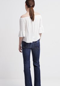 Top bianco off-the-shoulder con maniche a volant e tessuto testurizzato, abbinato a jeans a palazzo blu scuro con tasche posteriori.