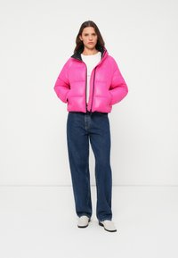 Veste doudoune rose vif avec fermeture éclair sur le devant, doublure intérieure noire et poignets élastiques, portée sur une chemise blanche et associée à un jean bleu.