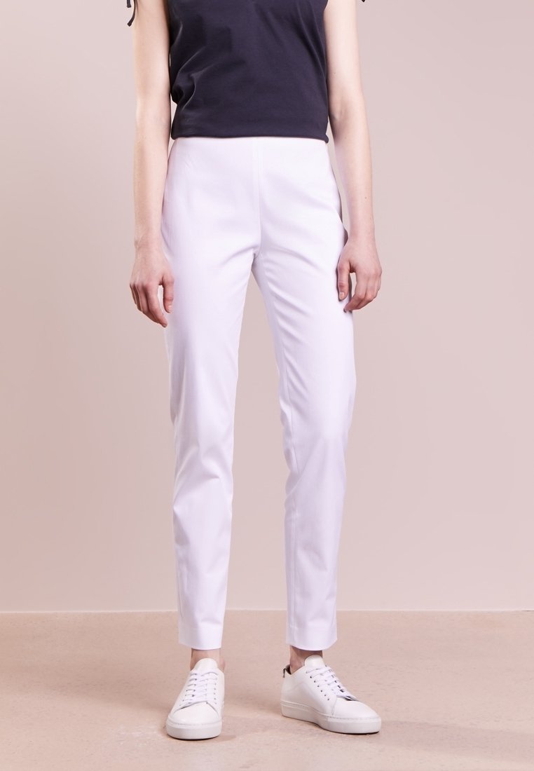 Pantalons blancs fuselés en tissu lisse, dotés d'une taille haute, de poches latérales, et associés à des baskets blanches sur un fond neutre.