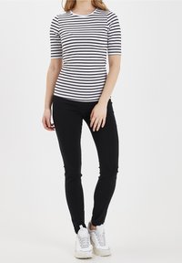 Kobieta ubrana w czarną i białą pasiastą bluzkę z krótkim rękawem, czarne wąskie spodnie i białe sneakersy na grubej podeszwie, stojąca na tle białego tła.