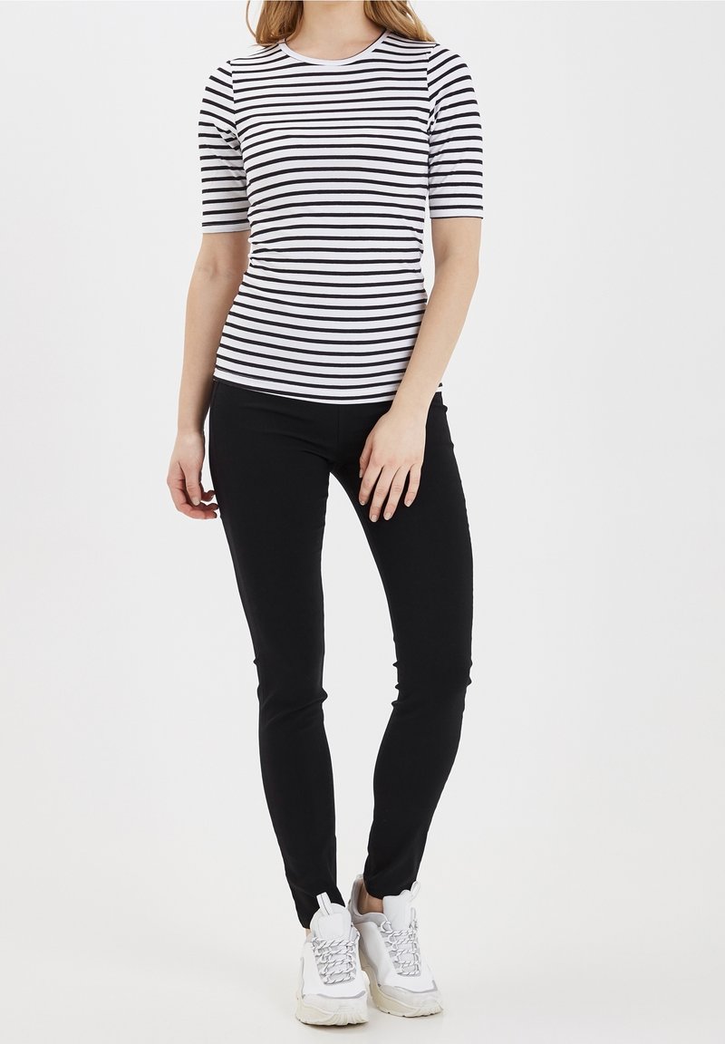 Kobieta ubrana w czarną i białą pasiastą bluzkę z krótkim rękawem, czarne wąskie spodnie i białe sneakersy na grubej podeszwie, stojąca na tle białego tła.