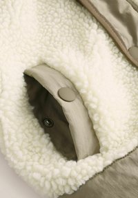 Beiges Außenmaterial mit cremiger Fleece-Auskleidung. Verfügt über eine Taschen mit Knopfverschluss und strukturierter Oberfläche. Weiche, gemütliche Textur mit kontrastierenden Farben.
