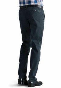 Jean en denim noir avec une coupe slim, présentant des poches arrière avec des fermetures à bouton et une ceinture en cuir. Porté avec des chaussures noires.