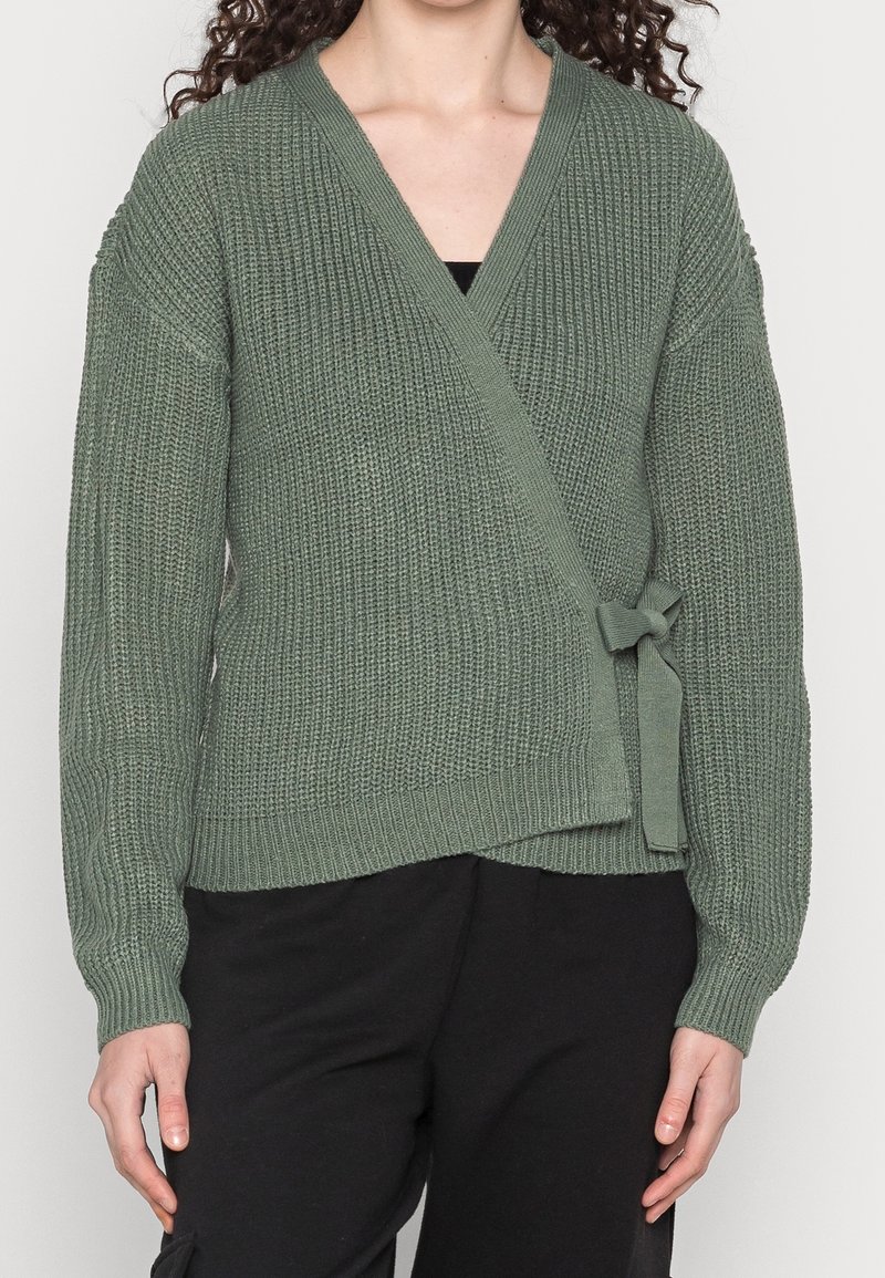 Femme portant un pull en maille vert sauge noué sur le côté, associé à un pantalon noir, debout devant un fond uni.