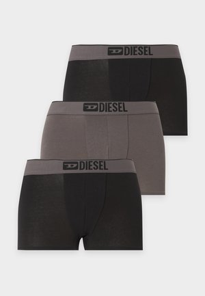 Jeune homme debout pieds nus portant un boxer Diesel noir sur fond blanc uni, regardant légèrement vers la gauche.