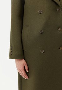 Dubbelknäppt olivgrön kappa i strukturerat tyg, med metallknappar och en sidoficka. Ärmslutet har fyra knappar.