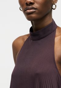 Κοντινό πλάνο μιας γυναίκας που φορά χρυσούς κρίκους στα αυτιά και αμάνικο μπλουζάκι με ψηλό γιακά σε σκούρο μωβ χρώμα, σε απλό φόντο.