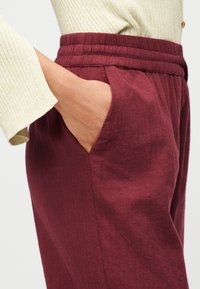 Χέρι στην τσέπη ψηλοκάβαλων, υφασμάτινων μπορντό παντελονιών σε συνδυασμό με κρεμ ριμπ μακρυμάνικο τοπ με φαρδύ τελείωμα.