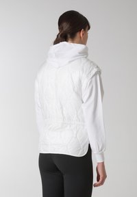 Gilet bianco trapuntato con vita elastica, colletto alto e maniche corte. Indossato sopra una maglietta bianca a maniche lunghe, abbinato a leggings neri.