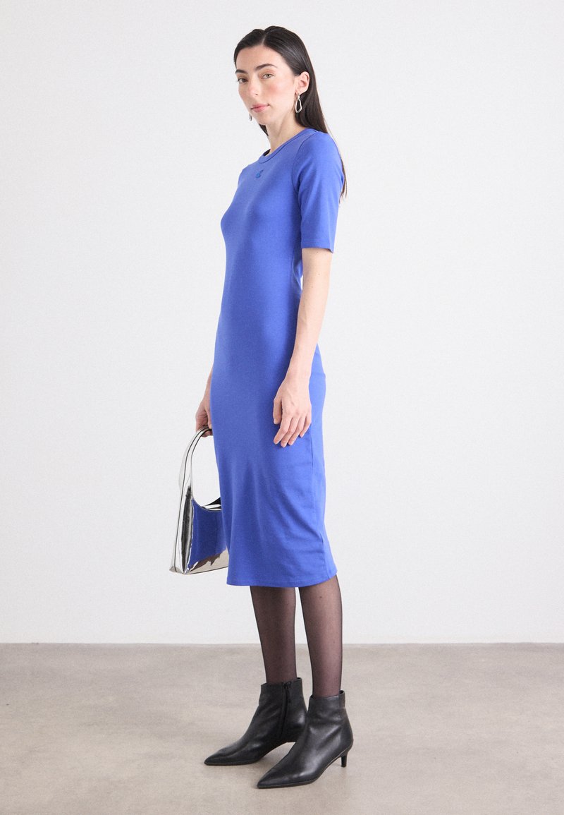 La modelo está de lado, vistiendo un vestido azul ajustado y botas negras hasta el tobillo, sosteniendo un bolso plateado brillante, en un ambiente minimalista interior.