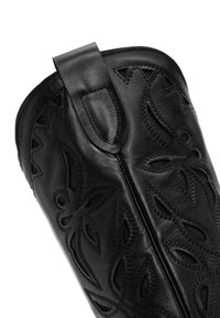Bota de cuero negro hasta la rodilla que presenta intrincados patrones florales troquelados, una textura suave y un lazo lateral para facilitar su uso.