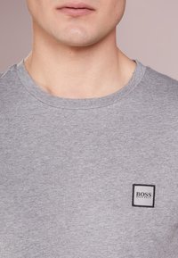 Šedé bavlněné tričko s kulatým výstřihem, které má malou čtvercovou nášivku s logem "BOSS" v černé barvě. Jednoduchá, hladká textura.