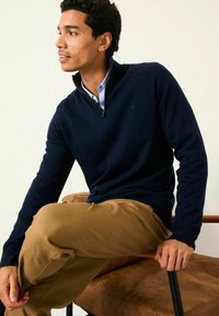 Navy Zip-Hals-Pullover aus weichem Material, mit geripptem Saum und Bündchen. Getragen über einem hellblauen Hemd mit Kragen und beigen Hosen.