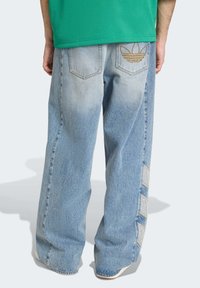 Slöa blå denimjeans med en blekt tvätt, med en guldfärgad Adidas-logotyp på bakfickan och kontrasterande detaljer på sidopanelerna.