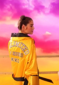 Mujer con el cabello recogido en un moño, vistiendo una chaqueta amarilla con "ILIMITADO 4 HORAS KIKO" en la parte posterior, contra un fondo vibrante de cielo rosa y naranja.