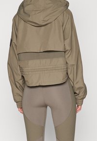 Chaqueta recortada de color verde oliva con capucha, que presenta una cintura ancha y puños elásticos, combinada con leggings ajustados en tonos apagados con acentos texturizados.