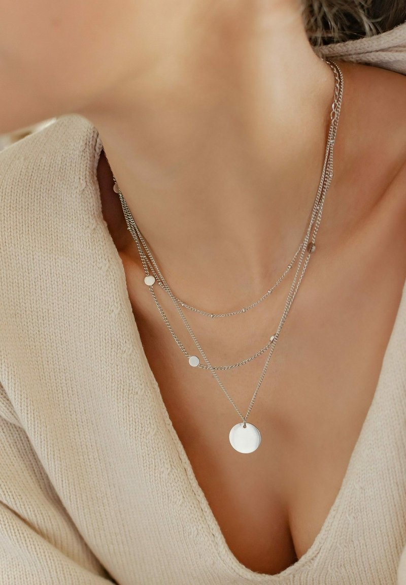Femme portant des colliers en argent superposés avec des pendentifs ronds et un pull beige en maille à col en V.
